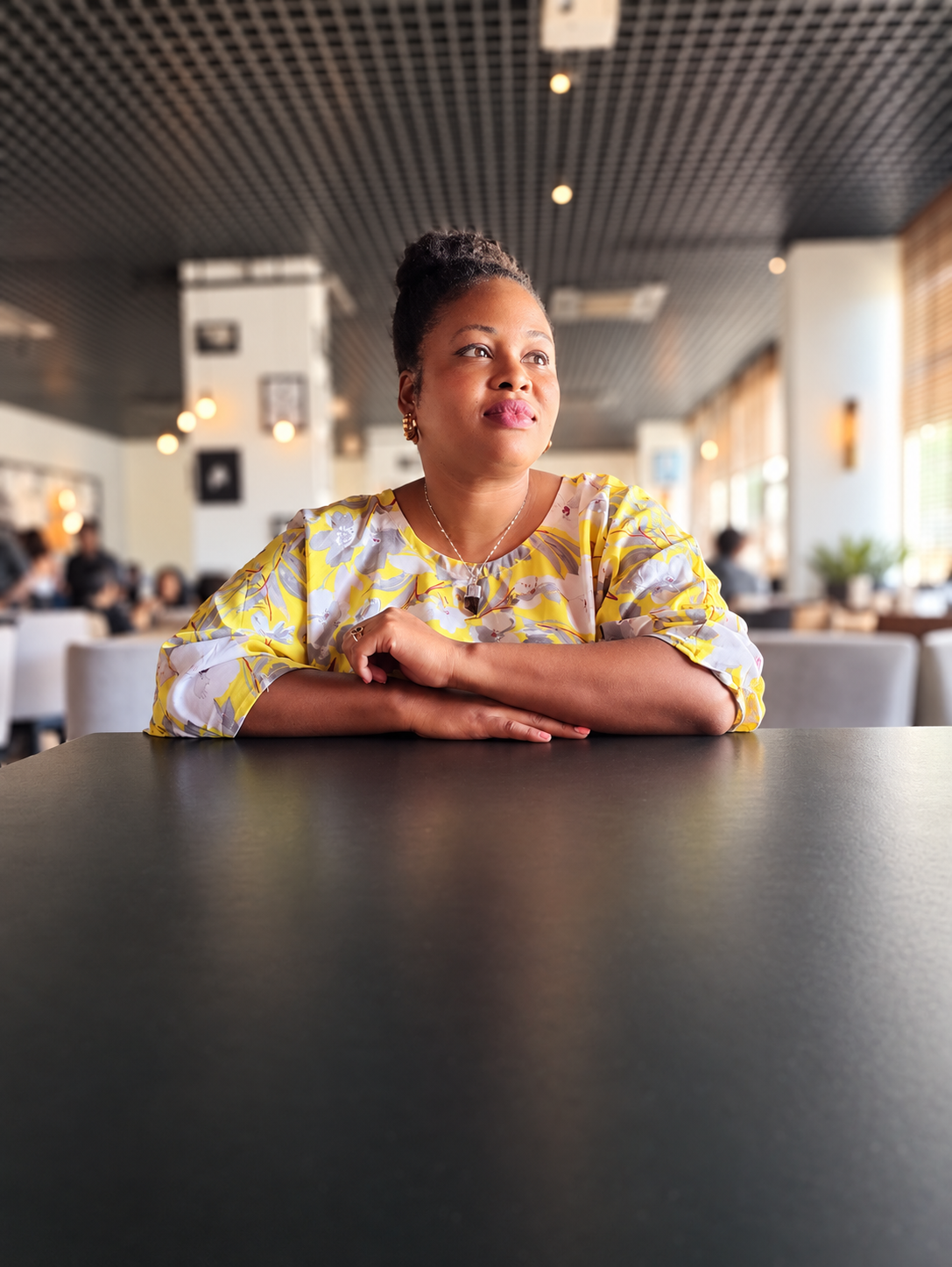 Tamara Ramsay seated at a café table in a yellow floral blouse, looking toward the light.